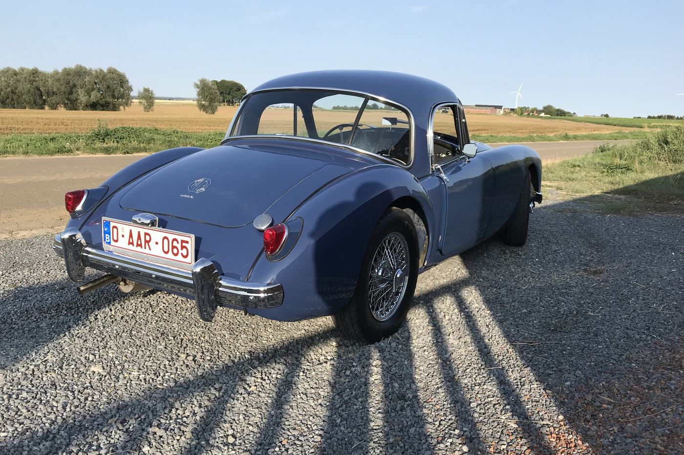 MGA coupé bleu de 1961 vue de trois quarts arrière sur un chemin de gravier, devant des champs